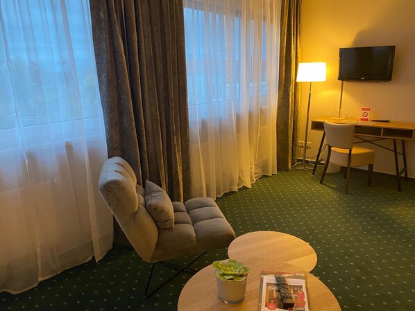 Une chambre d’hôtel comprenant un canapé, une petite table ronde avec une plante et des télécommandes, un bureau avec une chaise et un téléviseur fixé au mur.