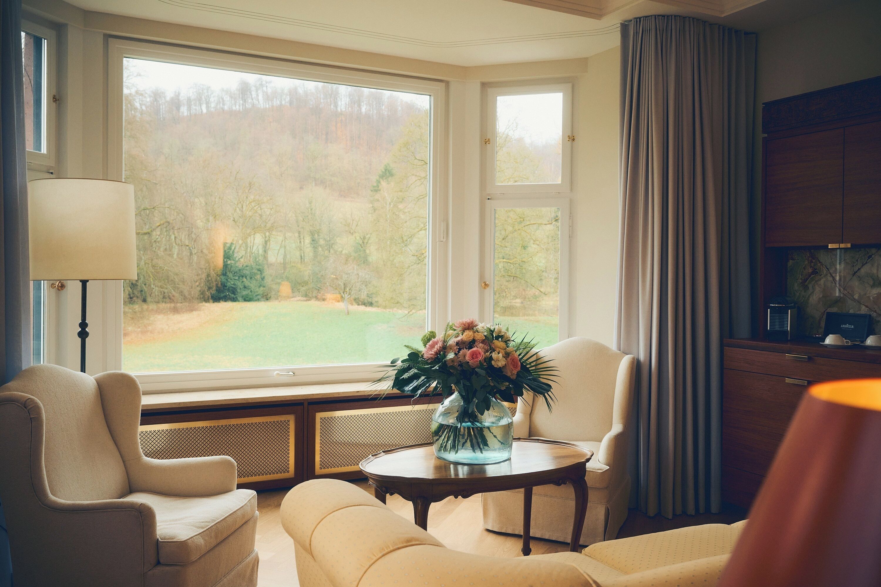A room with a large window, a vase with flowers, a lamp, and a wooden cabinet.