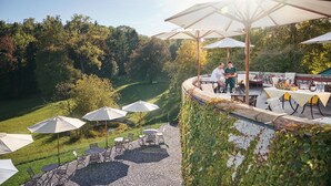Een buitenterras met tafels en stoelen, omgeven door groen en uitzicht op een aangelegde tuin.