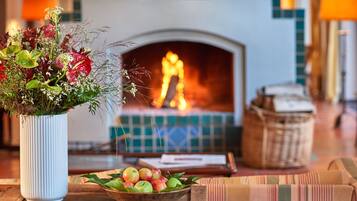 Una mesa de madera con un jarrón blanco de flores y un bol de manzanas. Al fondo, se ve una chimenea encendida.