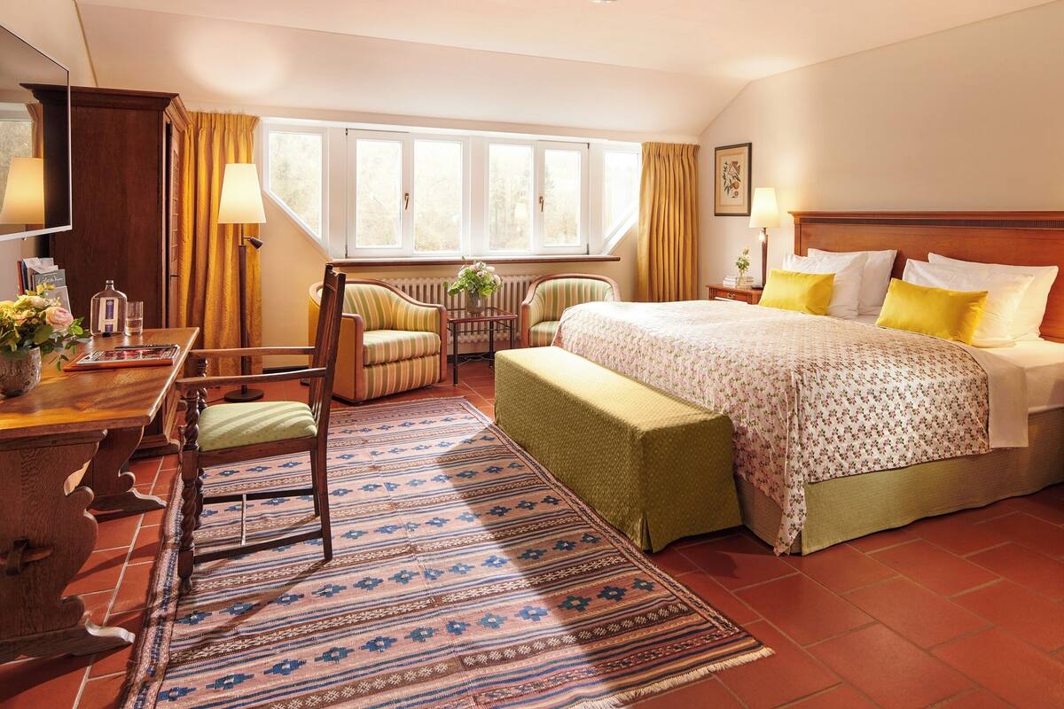 Una habitación de hotel con una cama, dos sillas, un escritorio, una ventana con cortinas, un radiador y una mesita de noche con una lámpara.
