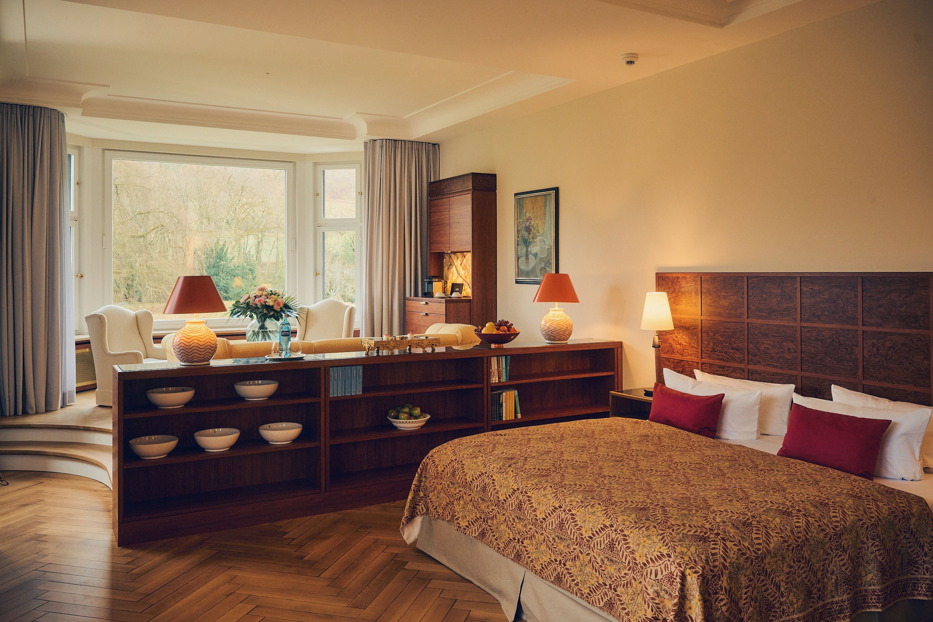 A hotel room with a bed, a desk with bowls, a chair, a lamp, and a painting on the wall.