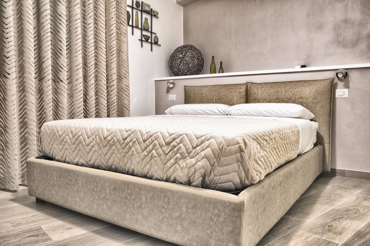 A bedroom with a large bed, a headboard, a bedside table, and a wall-mounted shelf with decorative items.