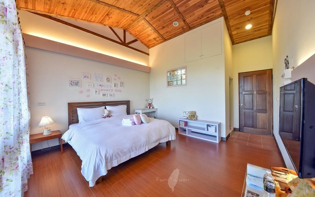 A bedroom with a wooden ceiling, a bed with white bedding, a nightstand with a lamp, a small table with a teddy bear, and a wardrobe.