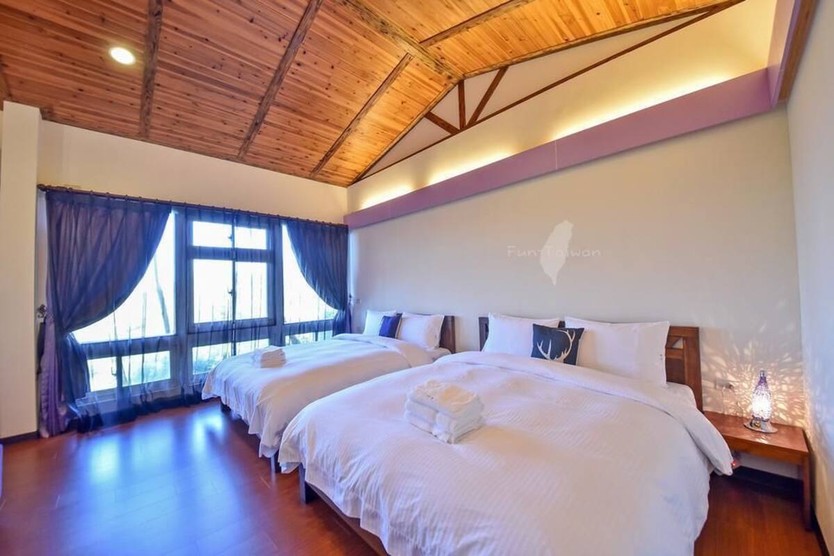 A hotel room with two beds, wooden ceiling beams, large windows with curtains, and a nightstand with a lamp.