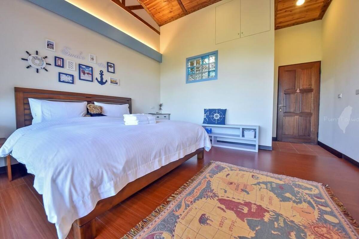 A bedroom with a large bed, wooden headboard, a nightstand, a bench, a wall clock, and a window with blue shutters.