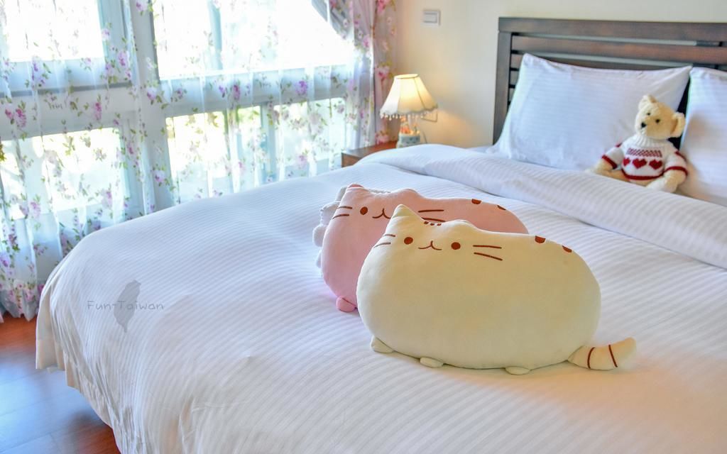 Cama bem arrumada com edredom branco e dois travesseiros macios em formato de gato. Há uma pequena mesa de cabeceira com um abajur e um ursinho de pelúcia sobre a cama.