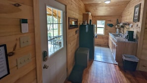 Interior de una cabaña de madera con una zona de cocina, una puerta con ventanal y un pequeño cubo de basura.