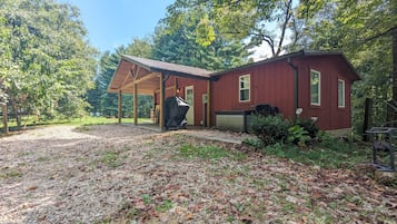 A red wooden house with a covered porch, surrounded by trees and a gravel driveway.