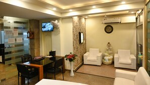 A hotel reception area with a reception desk, seating area with white chairs, a clock on the wall, and an air conditioning unit. - Ganga Darshanam Guest House (Varanasi)