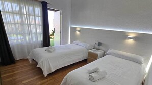 Habitación de hotel con dos camas, suelo de madera y vistas al exterior a través de cortinas de tul.