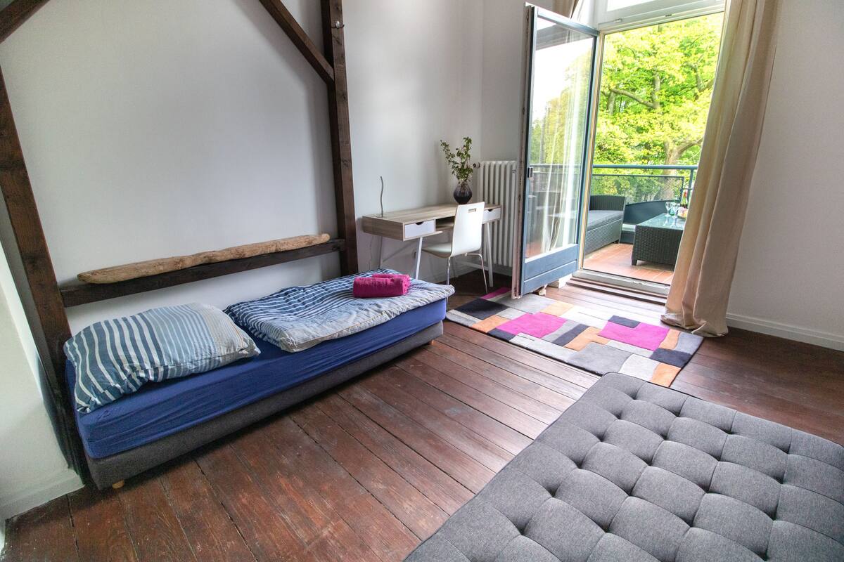 Habitación compacta con cama, escritorio y balcón con mobiliario exterior.