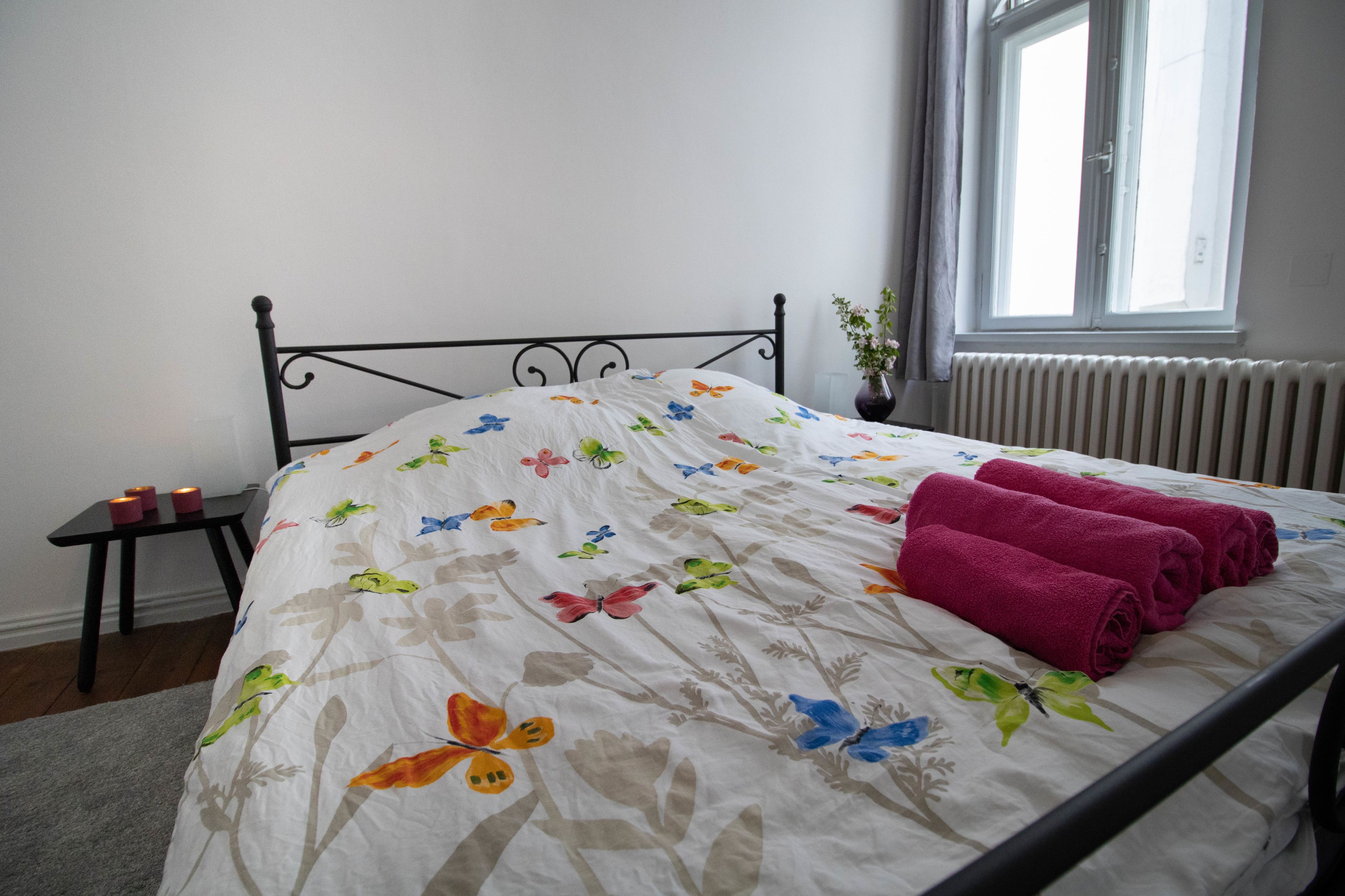 A bed with a floral bedspread and pink towels, a small table with candles, and a window with a view of the outside.
