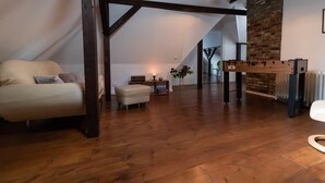 Un loft espacioso con techo alto, vigas de madera a la vista, un sofá acogedor, un pequeño reposapiés, una mesa de fútbol y una pared de ladrillo.