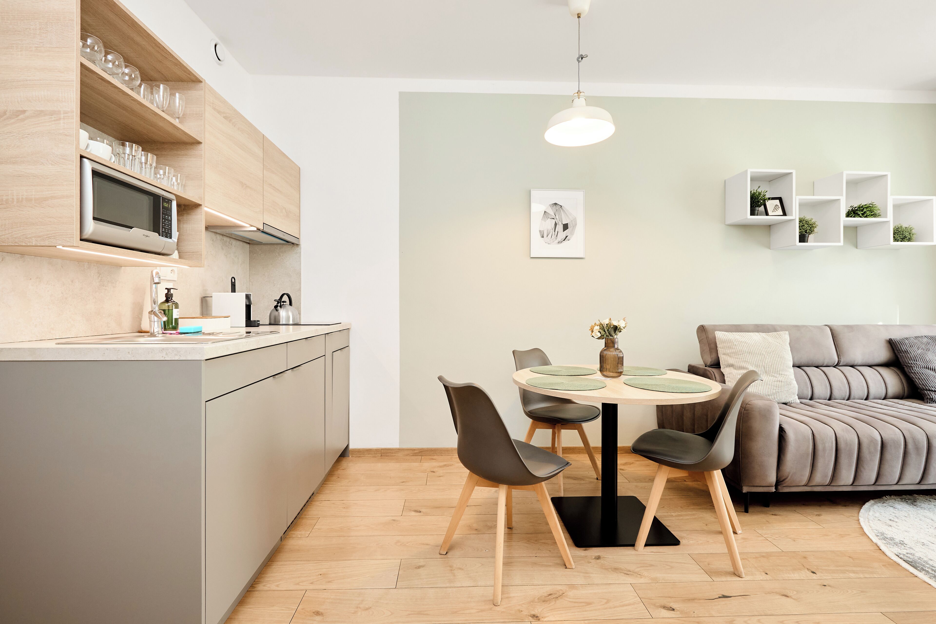 A modern kitchen with a microwave, sink, and wooden cabinets. There is a dining area with a round table and chairs, and a grey sofa in the living room.