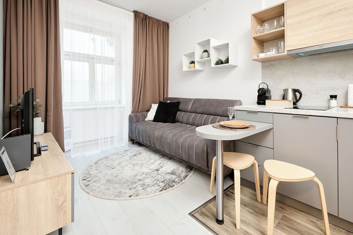 Una sala moderna con un sofá gris, una mesita redonda y dos taburetes de madera. Se observa un área de cocina con una encimera y una cafetera.