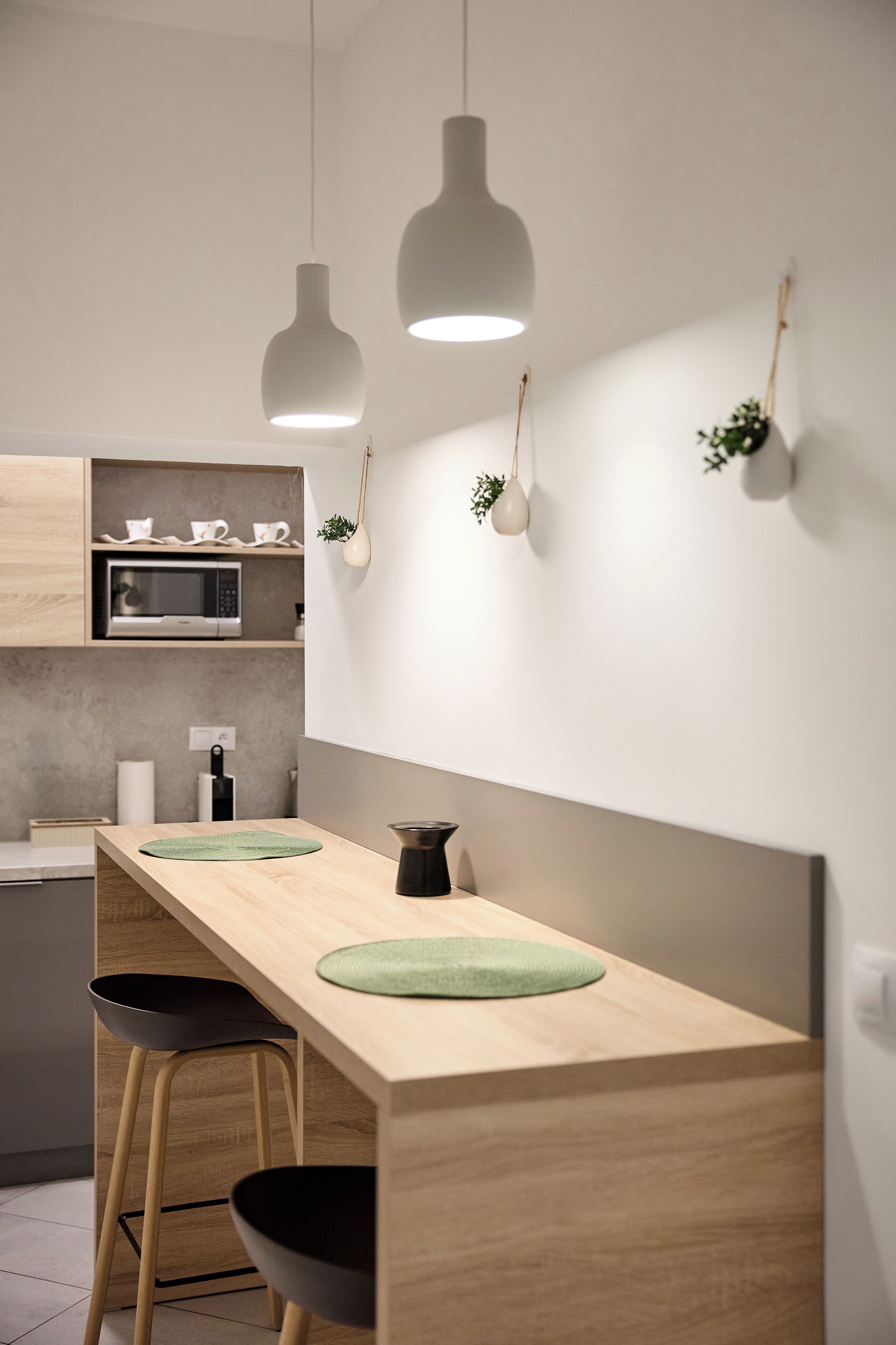 Eine moderne Küche mit Holzarbeitsplatte, zwei Barhocker, hängenden Pendelleuchten und einer Mikrowelle über einem Regal mit Tassen.