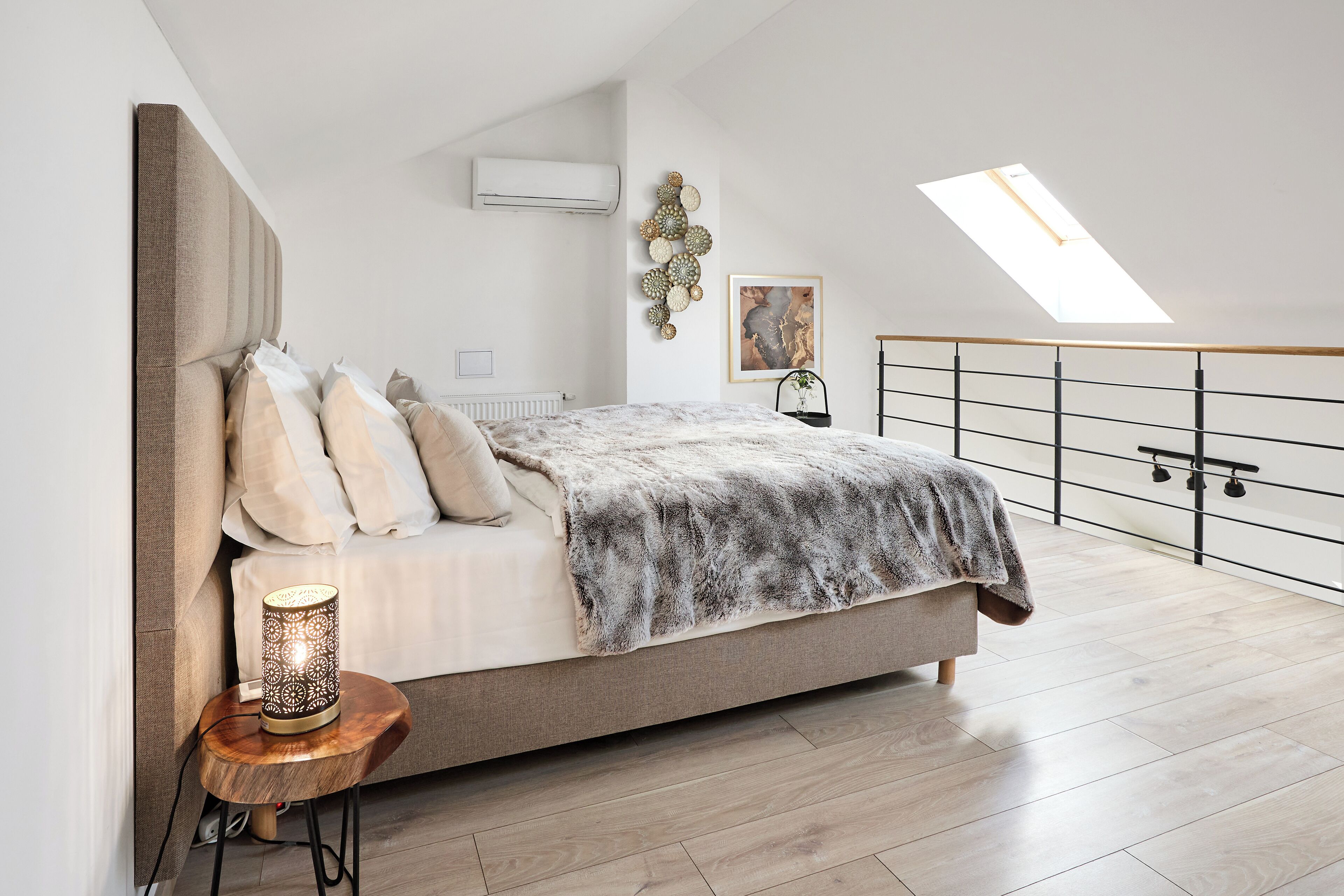 Een slaapkamer met een bed, een nachtkastje met een lamp, een radiator, een wandairconditioner, een decoratief wandobject en een dakraam.