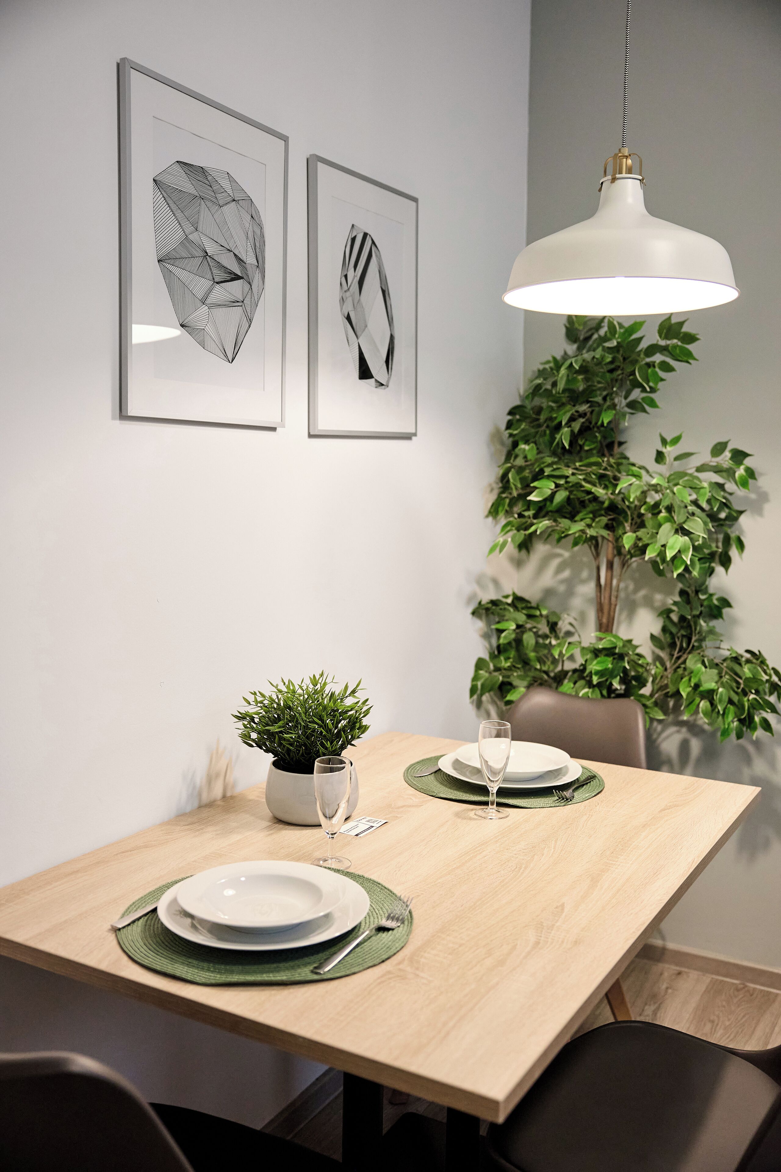 Una mesa de comedor con dos obras de arte geométricas enmarcadas, una lámpara colgante y una planta en maceta.