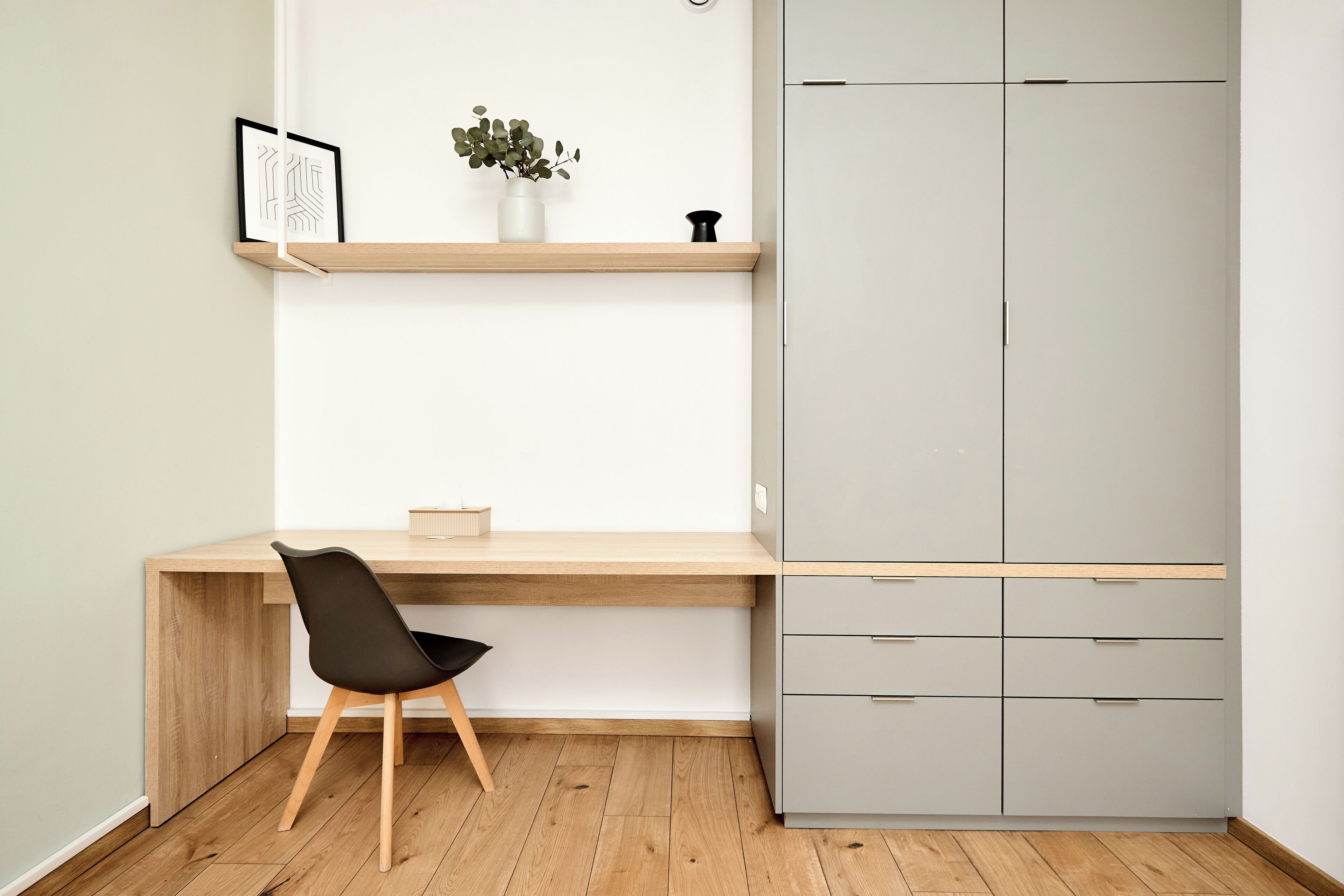 A modern room with a wooden desk, a black chair, a grey wardrobe, and a small shelf with a plant and a picture frame.