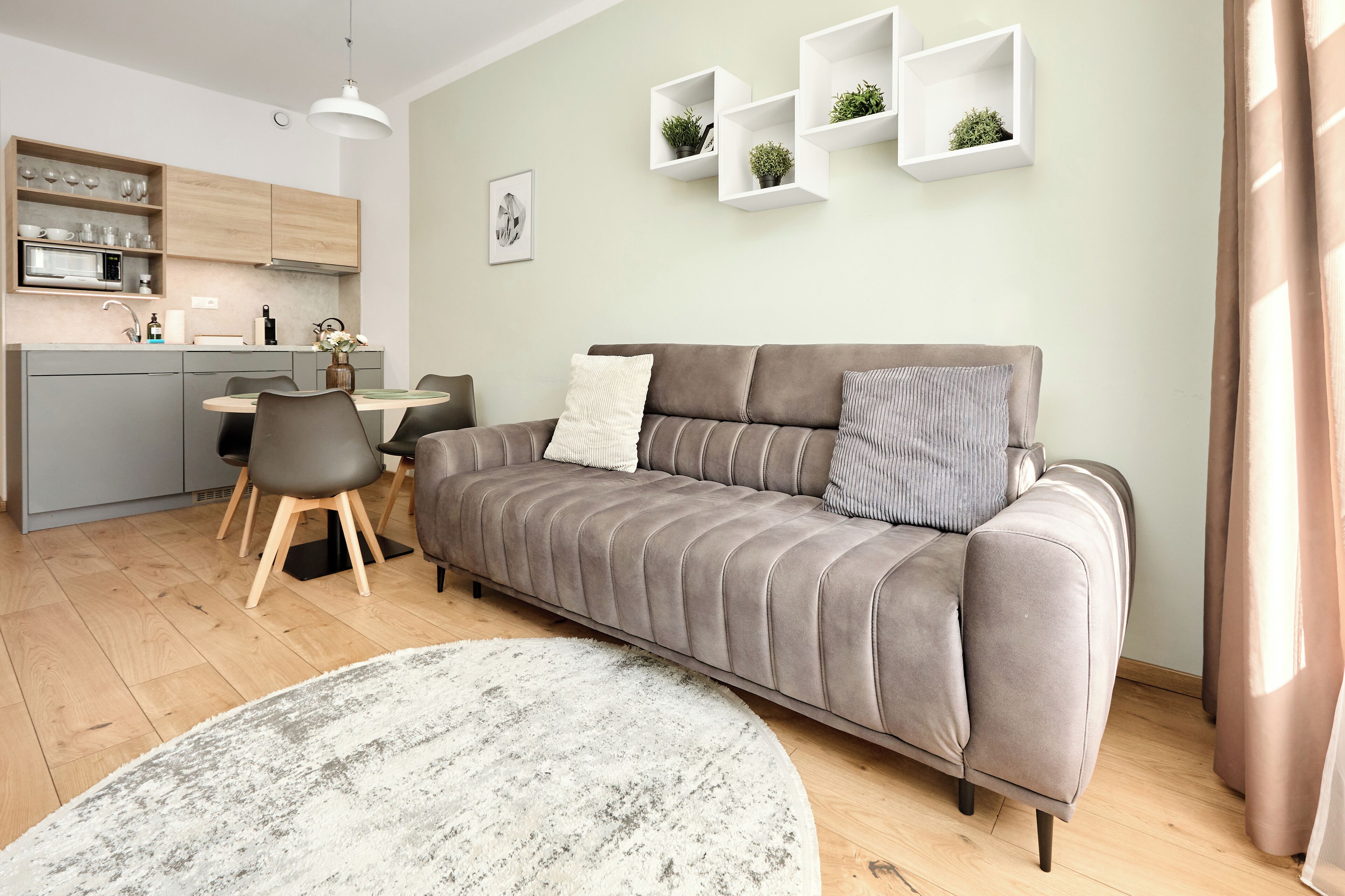 Een moderne woonkamer met een grijze bank, een eettafel met stoelen, een keuken en witte wandplanken met planten.