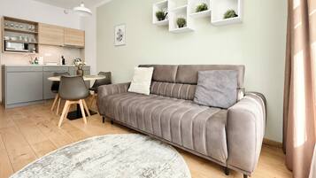 Una sala moderna con un sofá gris, una mesa de comedor con sillas, una zona de cocina y estanterías blancas con plantas.