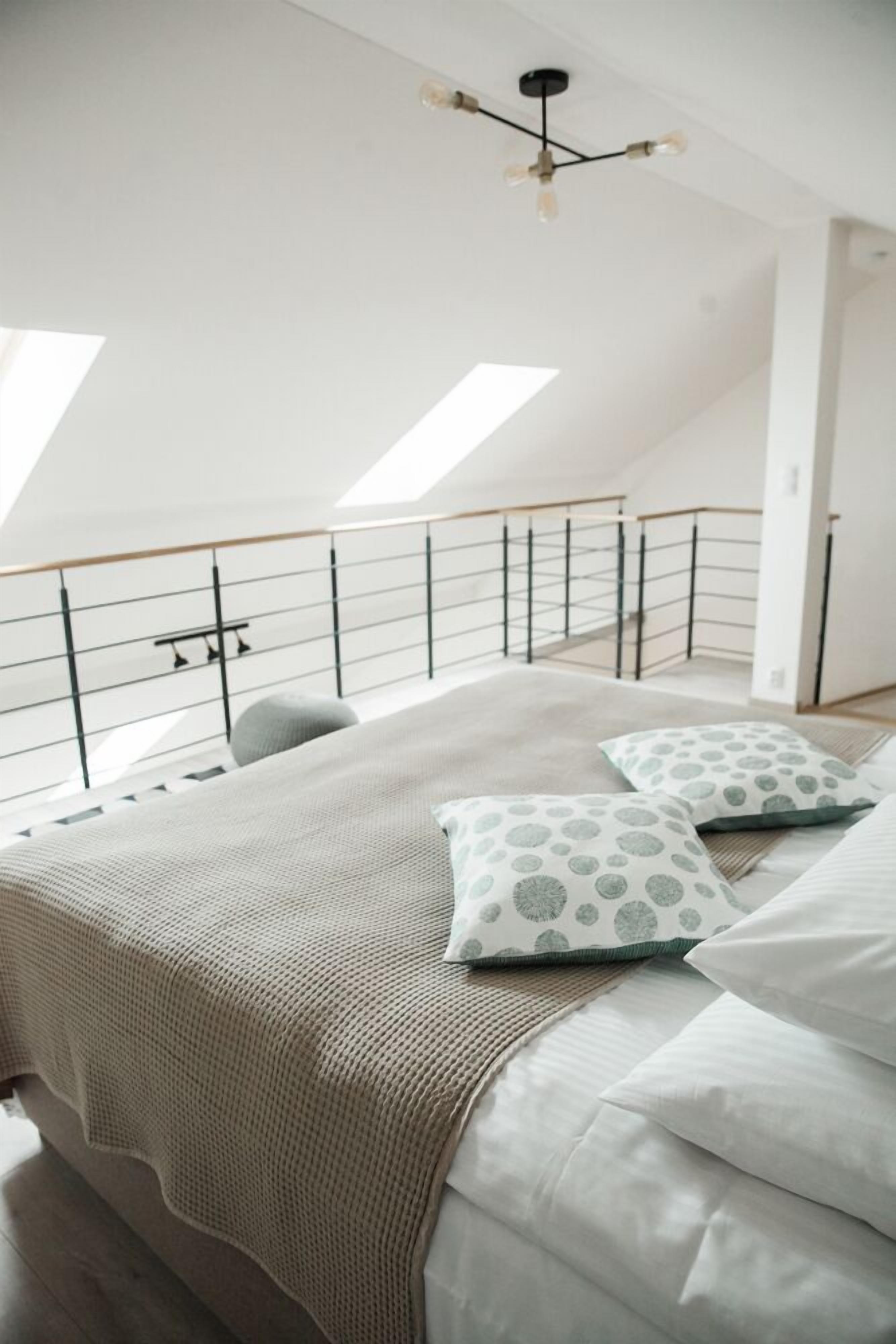 A bedroom with a high ceiling, skylights, a bed with a beige blanket, patterned pillows, and a modern chandelier.