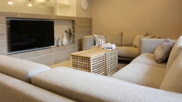 A modern living room with a large flat-screen TV mounted on the wall, a beige sofa, a coffee table, and recessed ceiling lighting. - The Neighbors (Osaka)