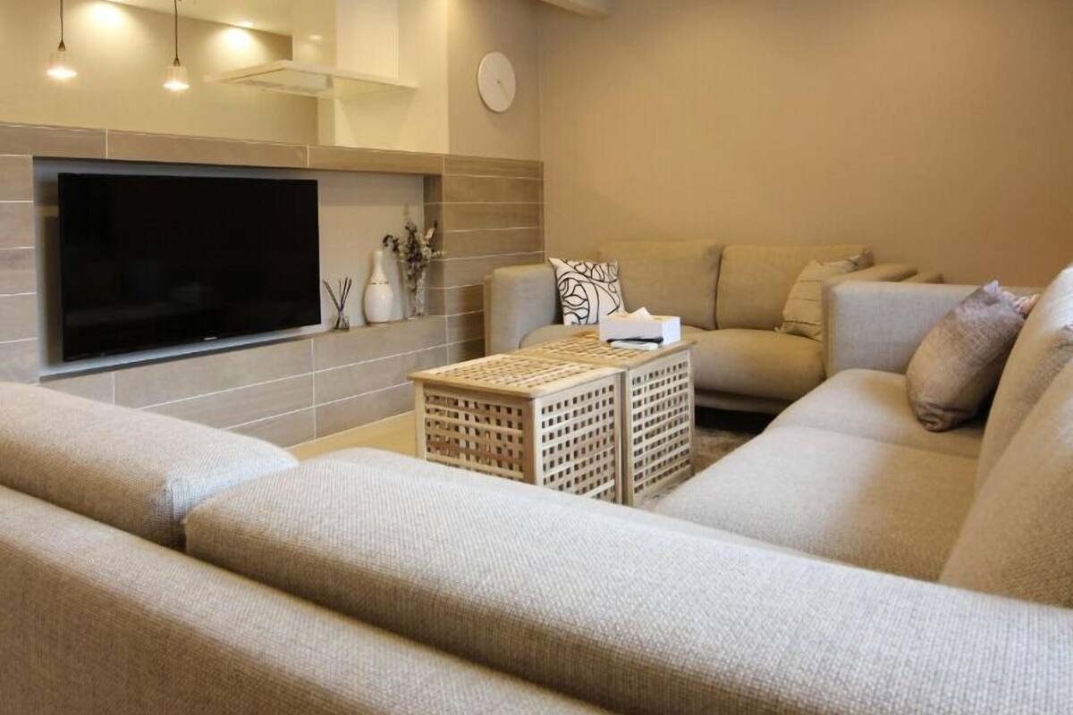 Una sala moderna con un televisor de pantalla plana grande montado en la pared, un sofá color beige, una mesa de centro e iluminación empotrada en el techo.
