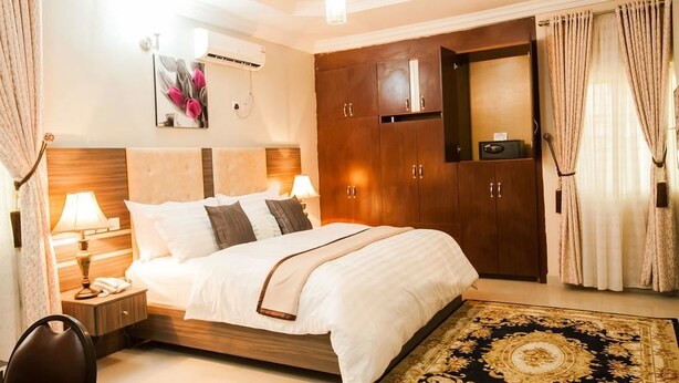 A hotel room with a large bed, wooden headboard, a nightstand with a lamp, a wardrobe, and a decorative rug. - Inti's Royal Suite (Abuja)