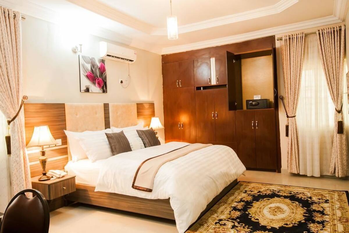 Une chambre d’hôtel avec un grand lit, une tête de lit en bois, une table de chevet avec une lampe, une armoire et un tapis décoratif.