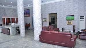 A lobby with stone columns, a reception desk, a red leather sofa, and a television mounted on the wall. - Kanem Suites (Abuja)