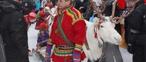 Une personne vêtue d’un costume traditionnel, accompagnée d’un renne, se tient au milieu d’une foule dans un décor extérieur enneigé.