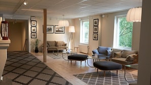 A modern living room with a sofa, ottoman, coffee table, and a floor lamp. There are framed pictures on the wall and a window with a view of trees. - Hotel Akerlund (Jokkmokk)