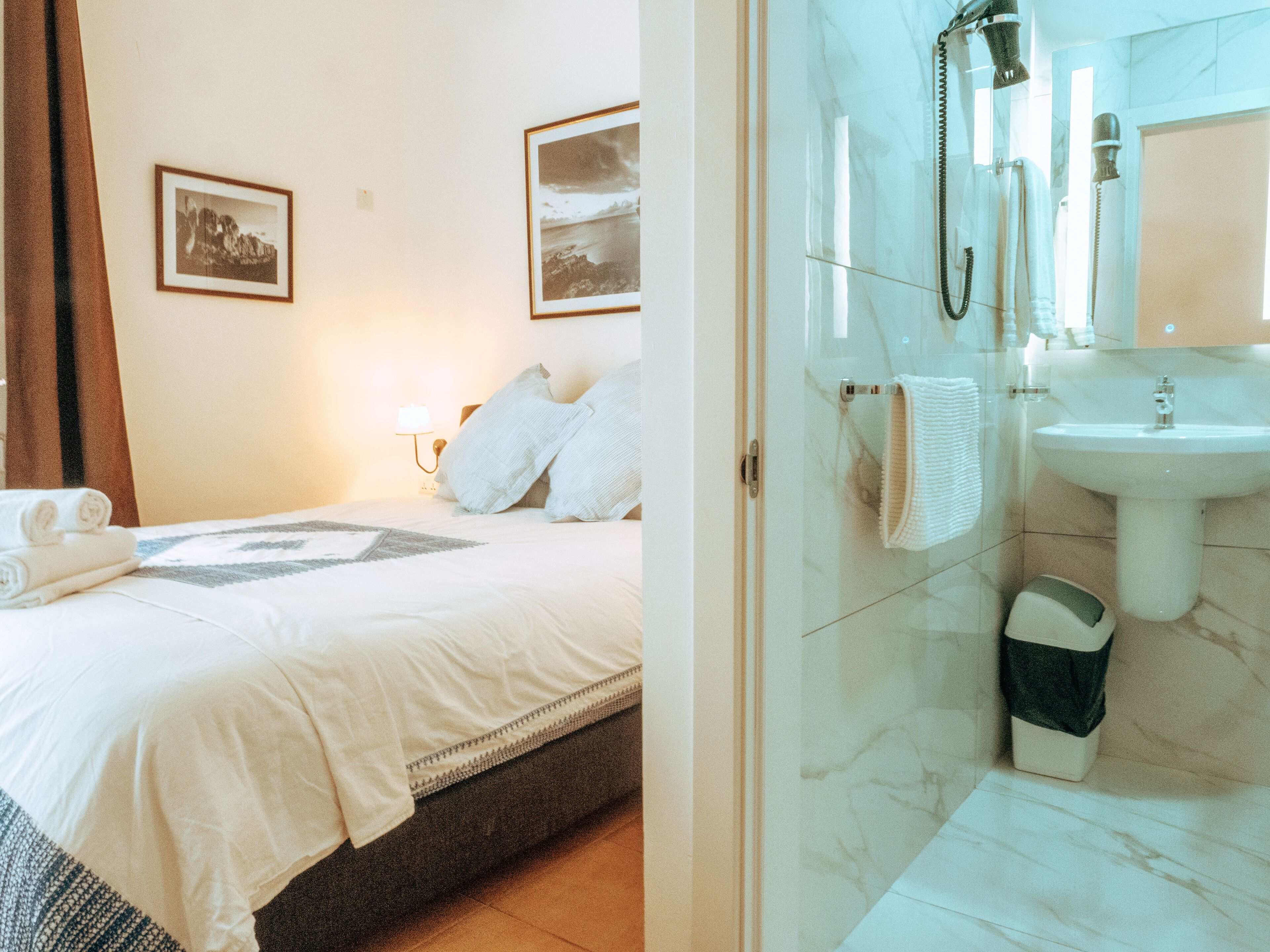 A hotel room with a bed, a bedside lamp, a framed picture on the wall, and a bathroom with a sink and shower.