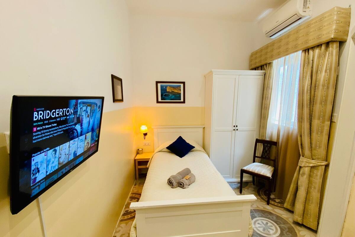A small hotel room with a bed, a TV mounted on the wall, a wardrobe, and a window with curtains.