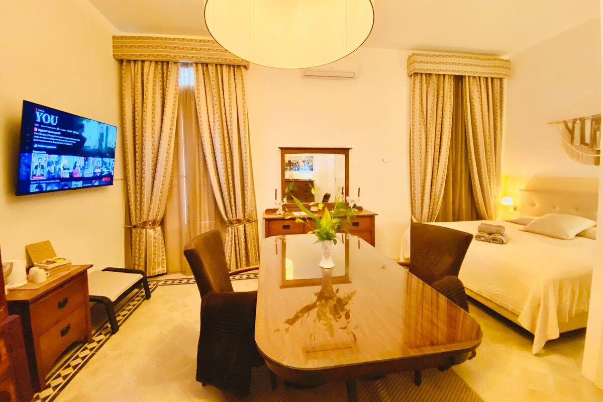 A hotel room with a wooden dining table, two chairs, a TV mounted on the wall, a bed with white bedding, and a large window with curtains.