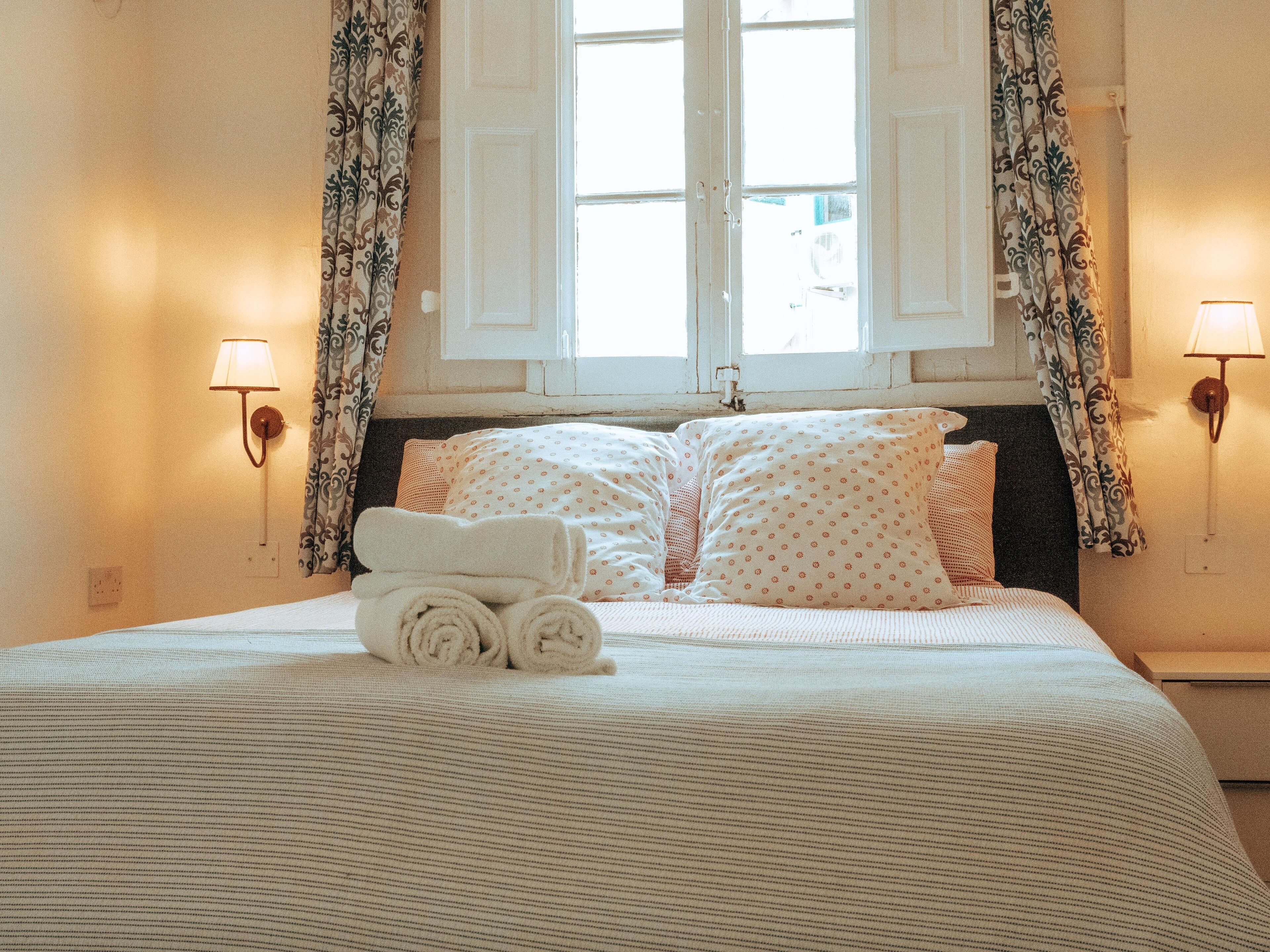 Una cama bien tendida con toallas enrolladas, un cojín estampado y una ventana con cortinas.