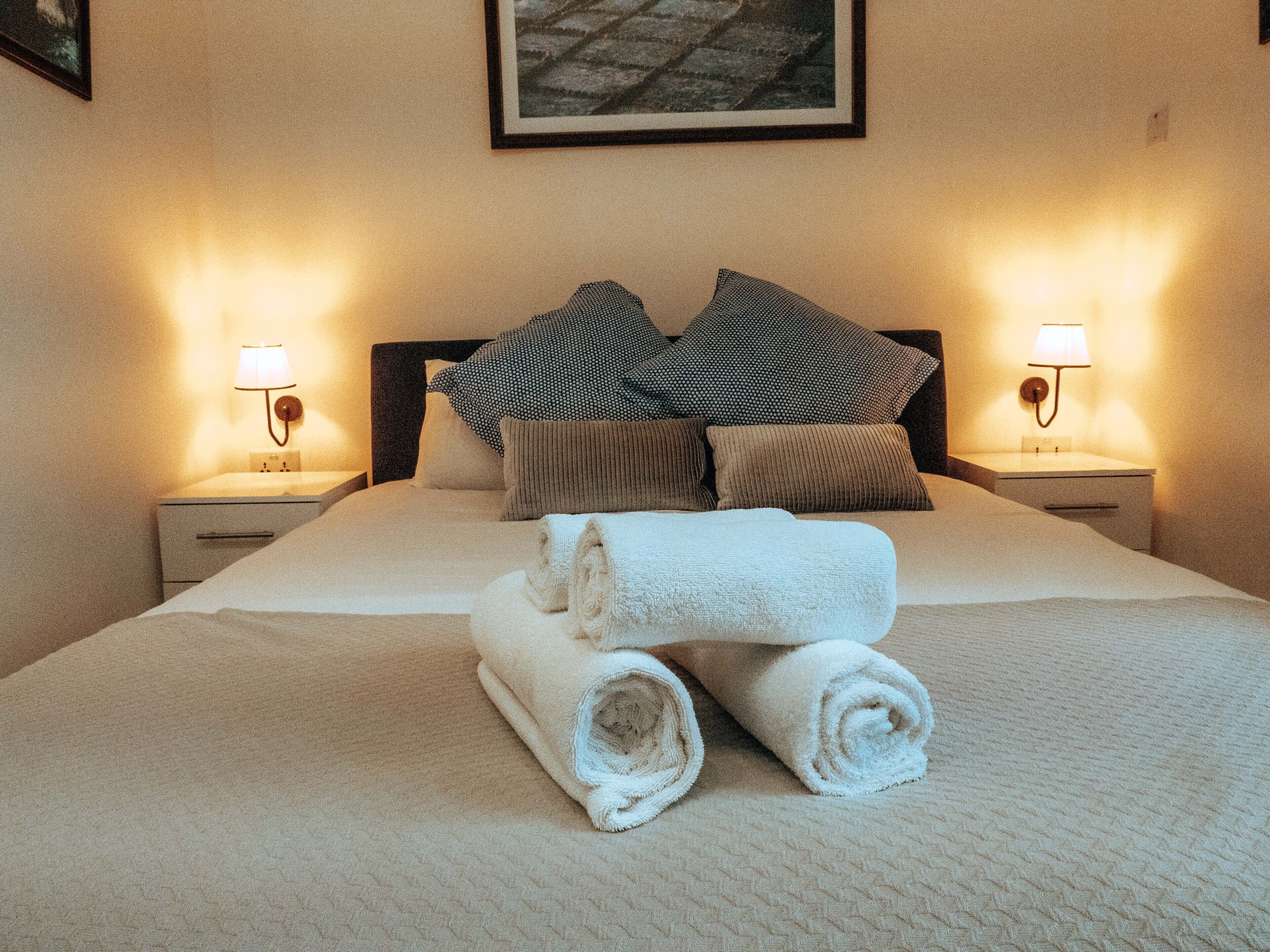 Una cama bien hecha con toallas enrolladas, dos mesitas de noche con lámparas y un cuadro enmarcado en la pared.
