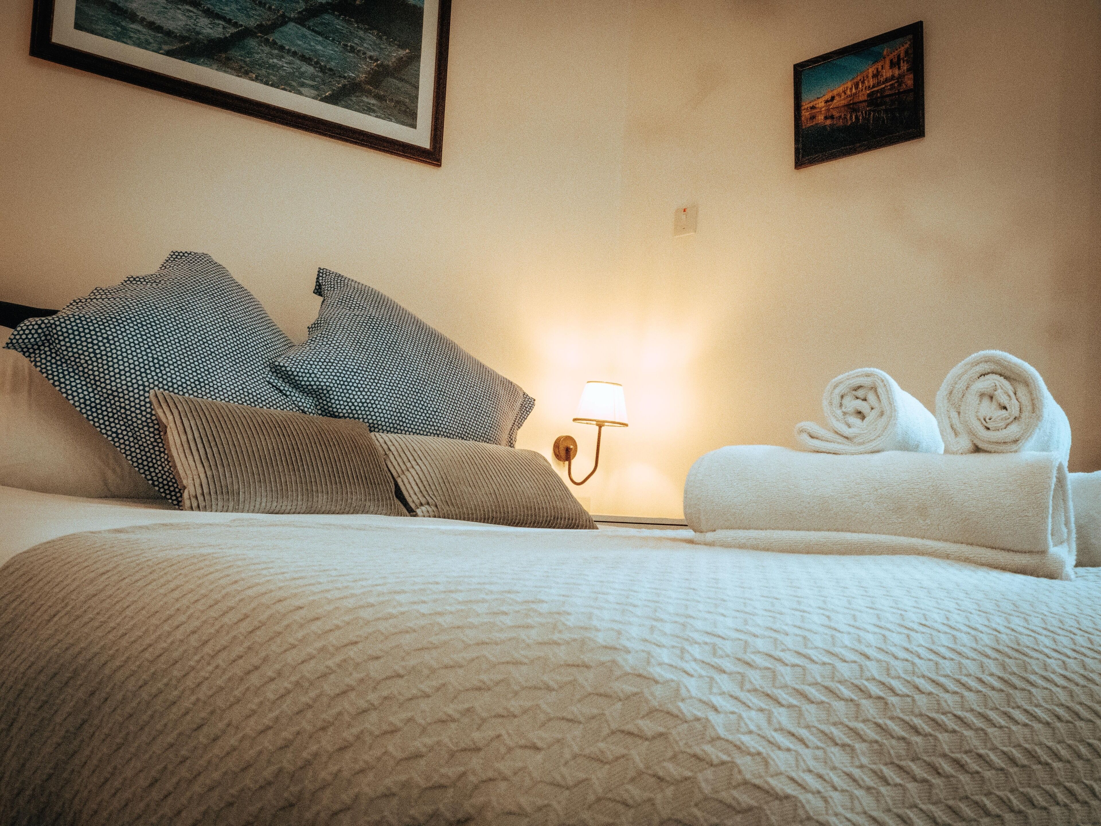 A neatly made bed with a textured white duvet, two rolled towels, and a wall-mounted lamp.