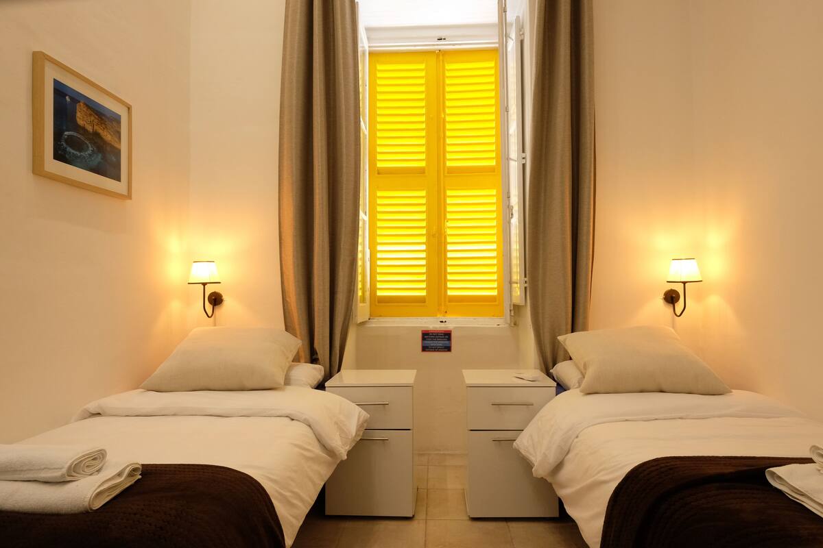 A hotel room with two beds, a window with yellow shutters, and wall-mounted lamps.