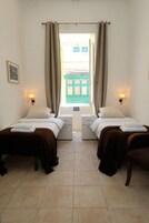 A room with two beds, each with a pillow and a blanket, a nightstand, a wall-mounted lamp, and a window with curtains.