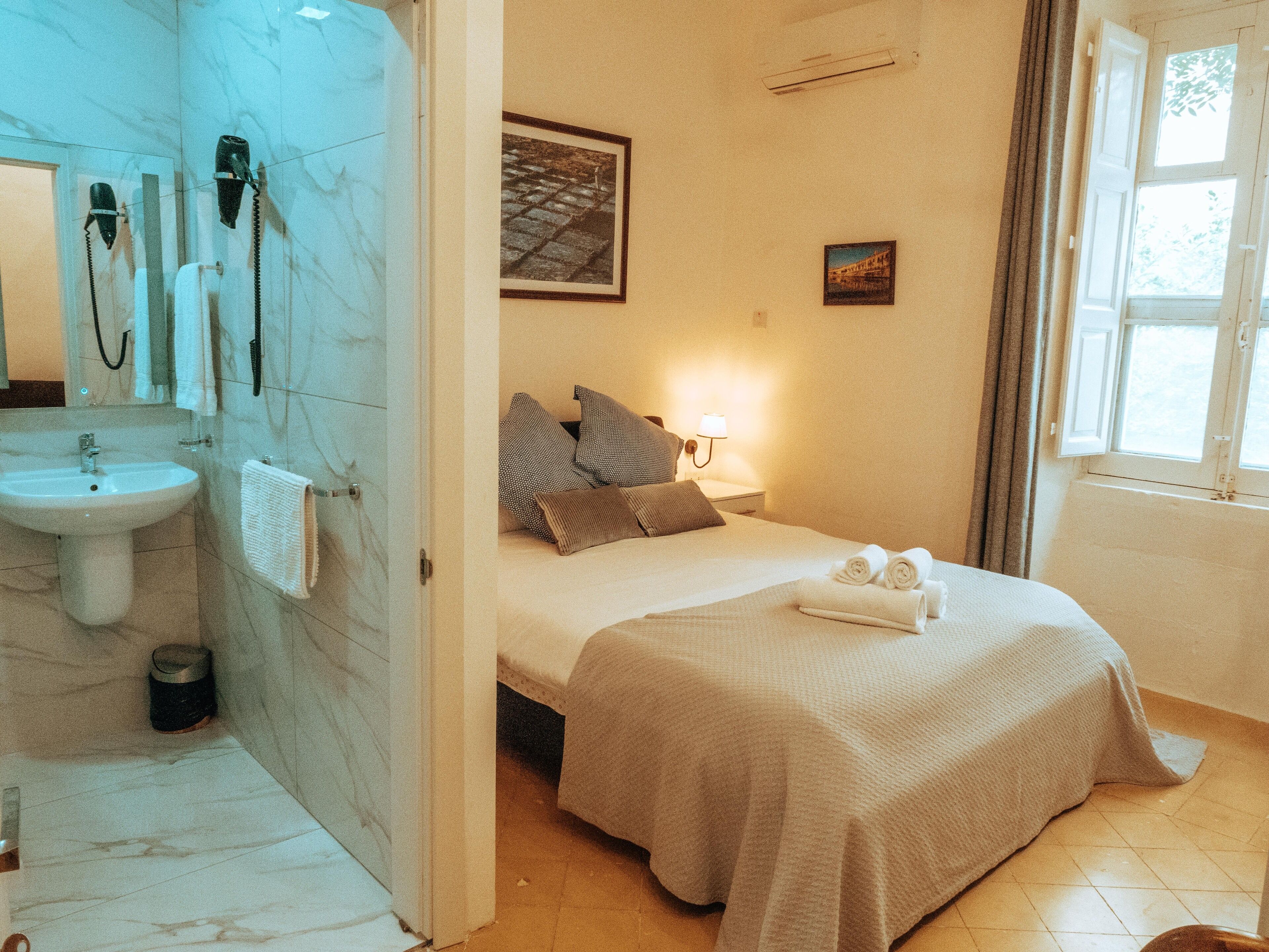 A hotel room with a bathroom, a bed with pillows, a bedside lamp, and a window.