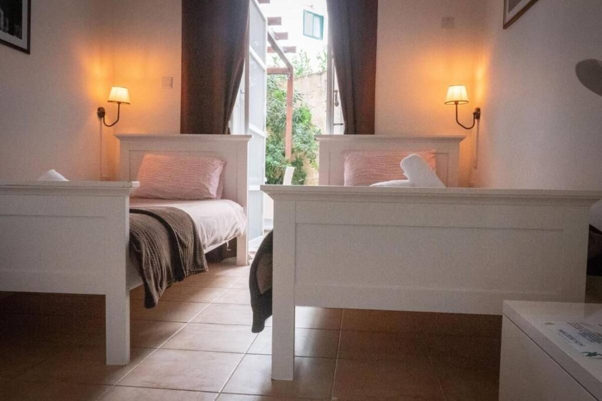 A hotel room with two beds, each with a headboard and a bedside table. There are two wall-mounted lamps on either side of the beds.