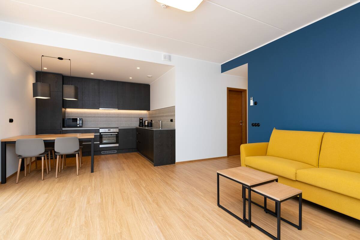 Eine moderne Küche mit dunklen Schränken, einem Holzesstisch und einem gelben Sofa in einem Wohnbereich mit blauer Akzentwand.