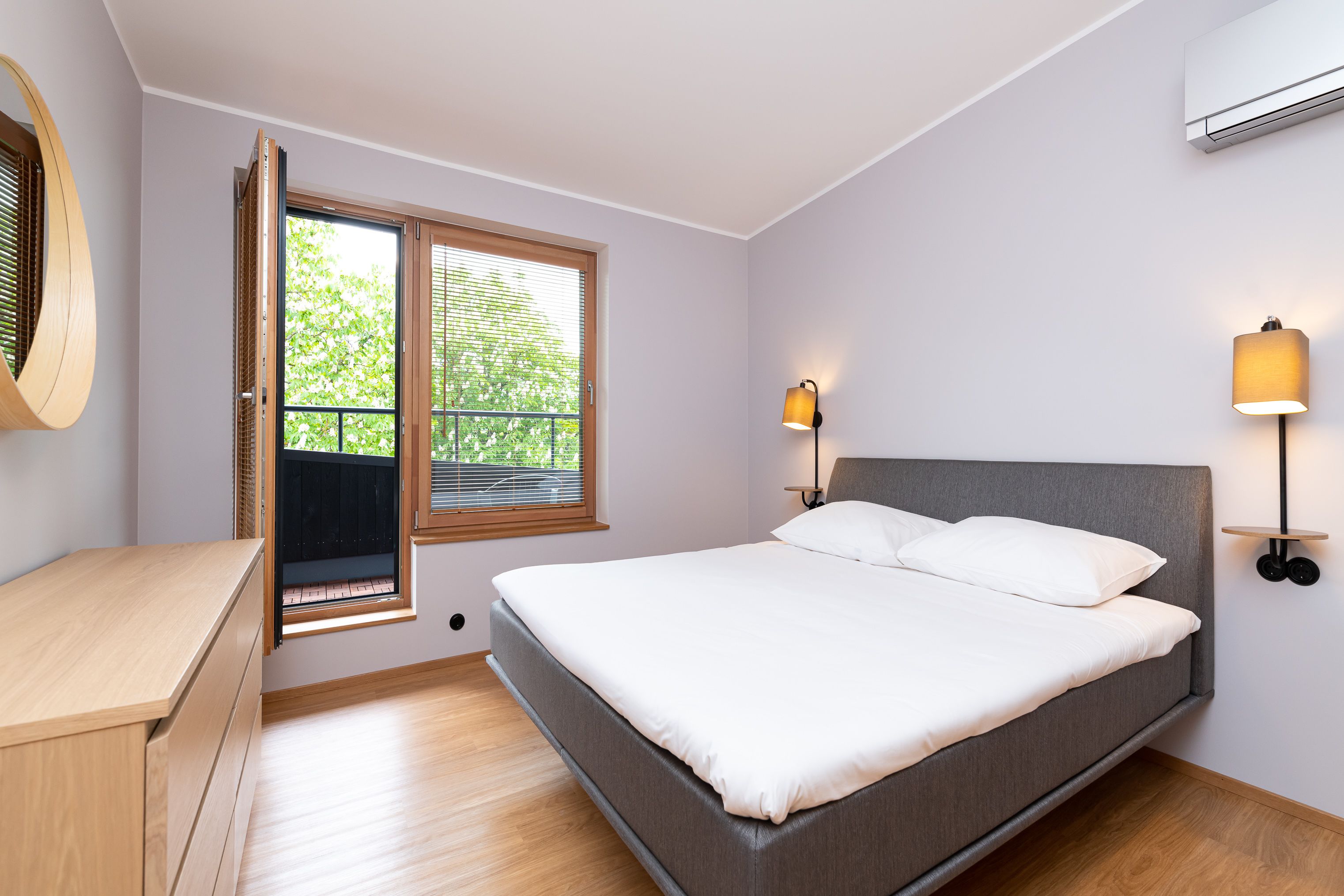 A bedroom with a bed, a wooden dresser, a window with blinds, and two wall-mounted lamps.