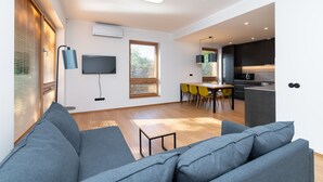 Uma sala de estar moderna com um sofá cinza, um espaço de refeições com cadeiras amarelas, uma cozinha com armários escuros e uma TV de tela plana fixada na parede.