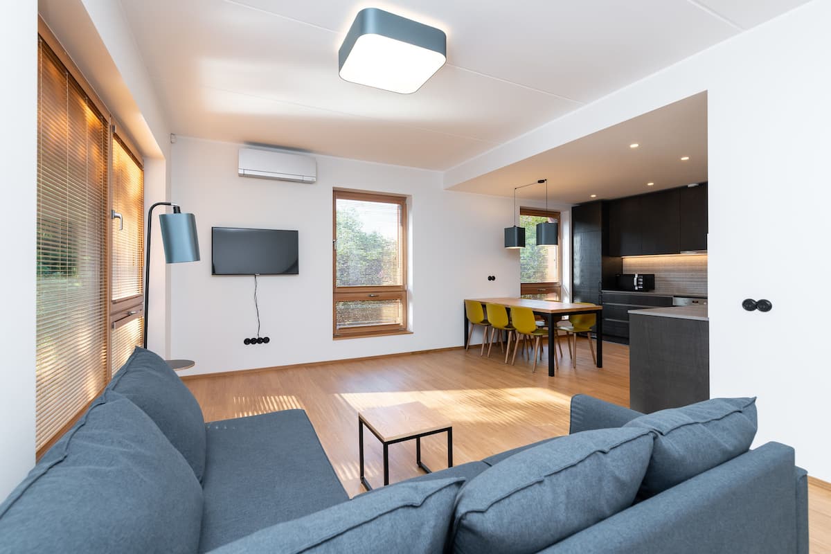 Ein modernes Wohnzimmer mit grauem Sofa, einem Essbereich mit gelben Stühlen, einer Küche mit dunklen Schränken und einem an der Wand befestigten Flachbildfernseher.