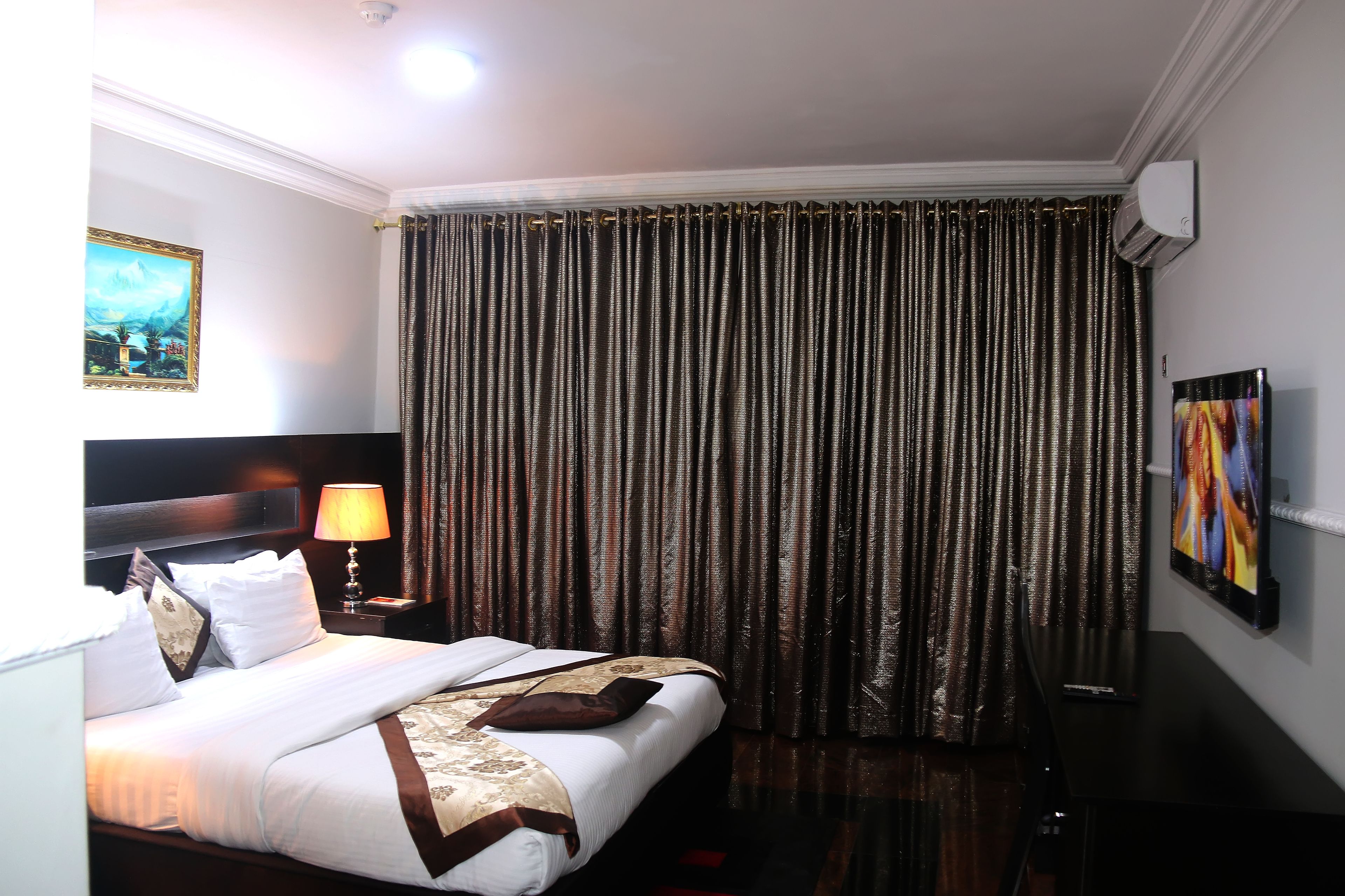 A hotel room with a neatly made bed, a bedside lamp, a painting on the wall, and a large window with curtains.