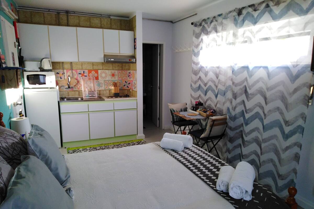 Habitación de hotel compacta con kitchenette, una cama con toallas enrolladas, zona de comedor y cortina con estampado en zigzag.